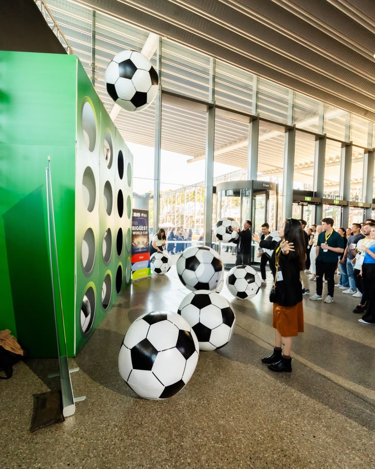 A convention entry hall with a tall green wall full of round pockets holding footballs and a school of giant inflatable soccer balls floating across the floor toward a woman in a brown skirt watching, with a screen behind reading 'BIGGEST WORLD CUP'.
