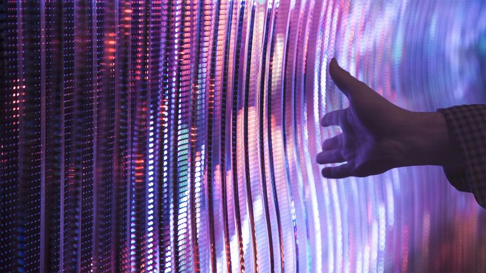 Curved LED wall made of thousands of bright reflective discs in purple, pink and white, a hand reaching out toward it from the right.