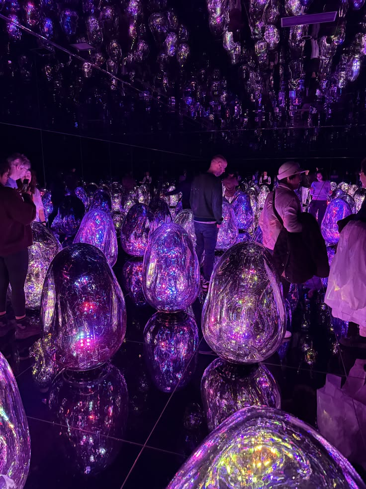 Immersive mirrored installation room: an infinite-mirror chamber filled with reflective ovoid sculptures glowing in iridescent purples, pinks and golds. Ceiling, walls and floor are mirrored, multiplying the sculptures and the visitors walking among them.