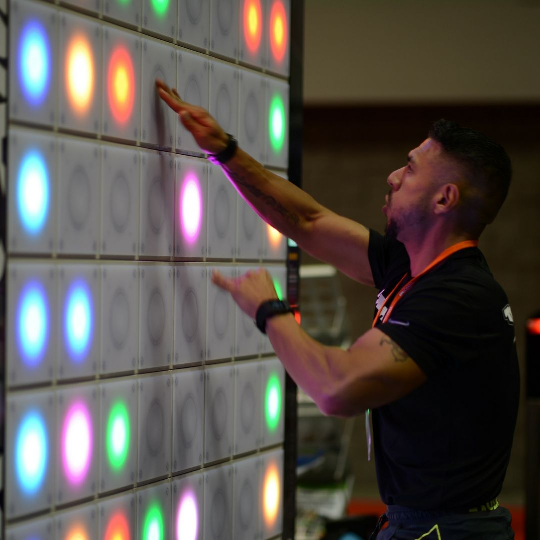 A person reaching up to touch a wall covered in a grid of square panels, each glowing with a single coloured LED disc (orange, pink, blue, green) inside a faint ring. A reach-and-tap interaction wall.